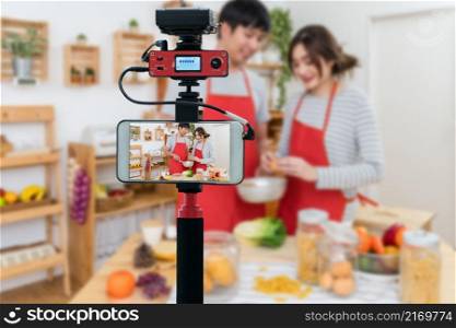 Closeup smart mobile phone taking Live of Happy Asian Lover or couple cooking in the kitchen room, Camera for photographer or Video and Live Streaming concept