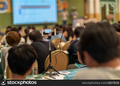Closeup smart mobile phone taking Live streaming with audience to Asian Speaker with casual suit standing and giving the knowledge on the stage in the conference hall or seminar meeting, business and education concept