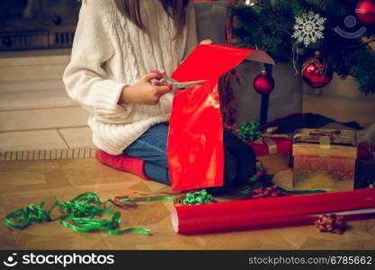 Closeup toned image of girl sitting under Christmas tree and cutting wrapping paper