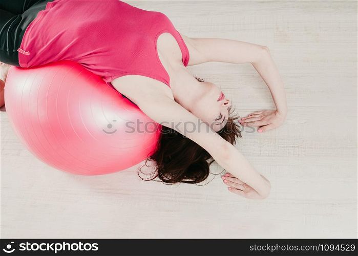 closeup top view of a young pretty woman in a pink top, making a bridge pose over a pink fitball in a gym, light floor background