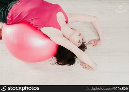 closeup top view of a young pretty woman in a pink top, making a bridge pose over a pink fitball in a gym, light floor background