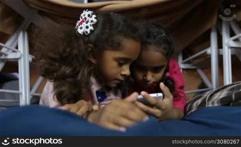 Closeup two adorable mixed race little sisters watching funny pictures on social network via smart phone as kids lie in cubby house made of chairs and blankets. Smiling african american girls playing online games on cellphone at home. Low view.