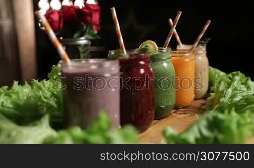 Closeup various freshly blended vegetable and fruit smoothies in mason jars with straws on wooden tray over bouquet of red roses and kitchen background. Dieting, vegetarian, fitness, healthy lifestyle.