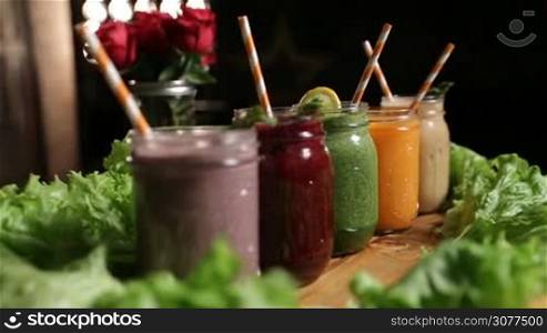 Closeup various freshly blended vegetable and fruit smoothies in mason jars with straws on wooden tray over bouquet of red roses and kitchen background. Dieting, vegetarian, fitness, healthy lifestyle.