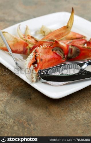 Closeup vertical photo of freshly cooked Dungeness crab legs on white plate with stainless crab crackers and stone counter top underneath