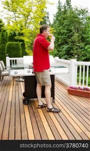 Closeup vertical photo of mature man drinking beer while looking at woods with BBQ grill and open cedar patio with seasonal trees in full bloom in background