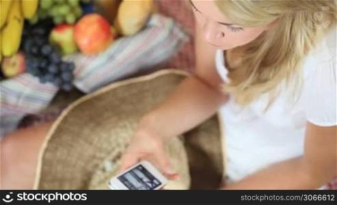 Closeup view of a beautiful blonde woman at a picnic siiting on a rug with fruit talking on her smartphone