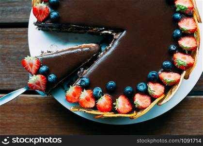 Closeup view of delicious chocolate cake decorated with fresh strawberries, blueberries, mint, and candied oranges on the brown wooden table. Luxurious dark glaze. Image for menu or confectionery catalog