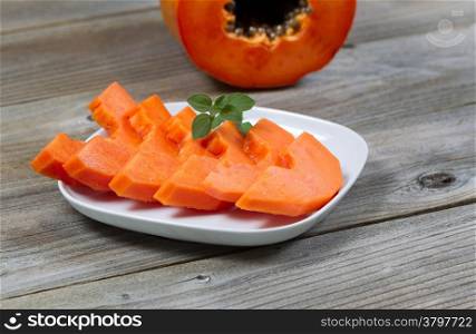 Closeup view of sliced papaya fruit, on white plate, with mint leaf and half piece in background on rustic wood