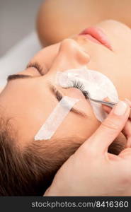 Closeup view of young caucasian woman having eyelash extension procedure in beauty salon. Beautician glues eyelashes with tweezers. Woman having eyelash extension procedure