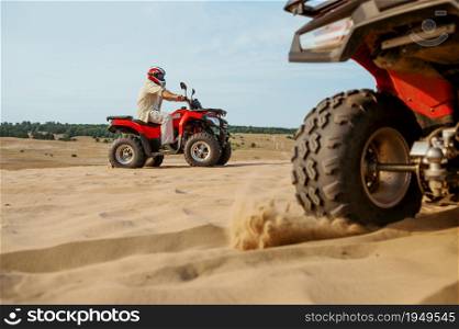 Closeup view on atv wheel, man in helmet on background. Male person on quad bike, sandy race, dune safari in hot sunny day, 4x4 extreme adventure, quad-biking. View on atv wheel, man in helmet on background