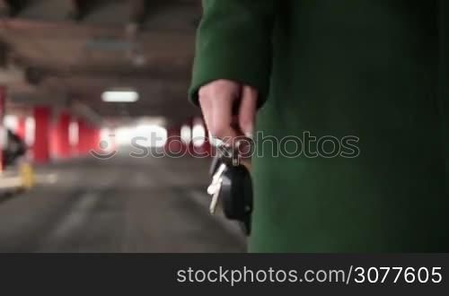 Closeup woman&acute;s hand holding car key with remote control car alarm system with blur vehicle parking lot while walking in underground garage. Middle section of woman walking in covered parking garage driveway with car key remote control in hand. Slow motion.