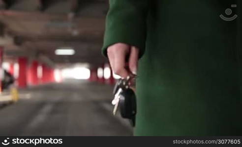 Closeup woman&acute;s hand holding car key with remote control car alarm system with blur vehicle parking lot while walking in underground garage. Middle section of woman walking in covered parking garage driveway with car key remote control in hand. Slow motion.