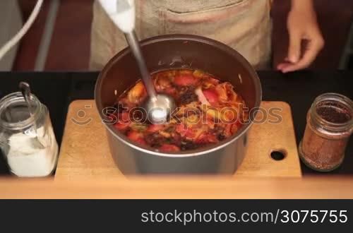 Closeup woman&acute;s hand mixing vegetable soup with blender in black pot on wooden board