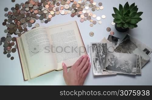 Closeup woman&acute;s hand turning page of vintage book. Old coins of different countries and vintage photos and postcards on the white table