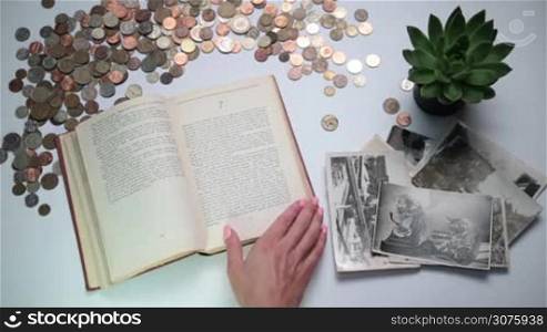 Closeup woman&acute;s hand turning page of vintage book. Old coins of different countries and vintage photos and postcards on the white table