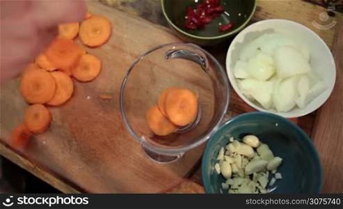 Closeup woman hands adding sliced carrot into a glass bowl. Chopped garlic,onion,red pepper in ceramic bowl on the table. Top view