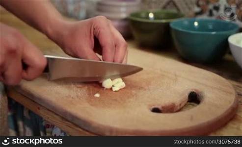 Closeup woman hands chopping fresh garlic with knife on cutting board .