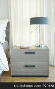 Closeup wooden night table with reading lamp, coffee cup and book in modern bedroom