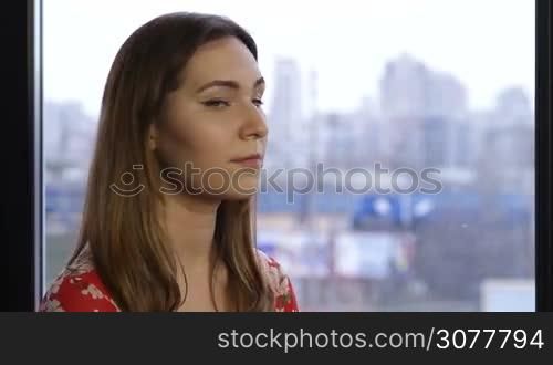 Closeup young attractive woman with clean skin and natural make-up standing at the window and smiling. Beauty woman with white perfect smile looking at camera in domestic interior over cityscape background. Positive emotion and facial expression.