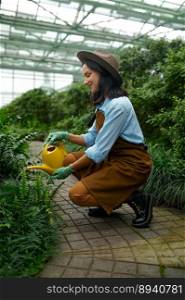 Closeup young woman florist gardener with watering can caring for plants pouring water into flowerbed in hothouse. Young woman gardener with watering can caring for plants in hothouse