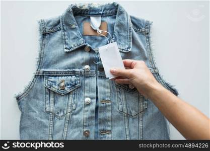 clothes, wear and fashion concept - close up of hand holding price tag of denim waistcoat