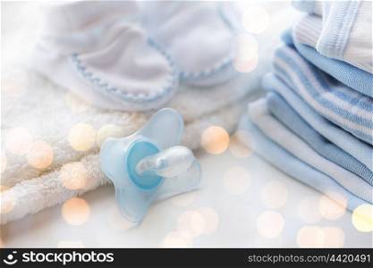 clothing, babyhood, motherhood and object concept - close up of baby soother, bootees and pile of clothes for newborn boy with holidays lights
