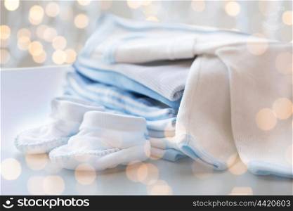 clothing, babyhood, motherhood and object concept - close up of white baby cardigan with bootees and pile of clothes for newborn boy with holidays lights