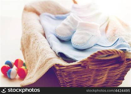 clothing, babyhood, motherhood and object concept - close up of white baby bootees with pile of clothes, towel and rattle for newborn boy in basket