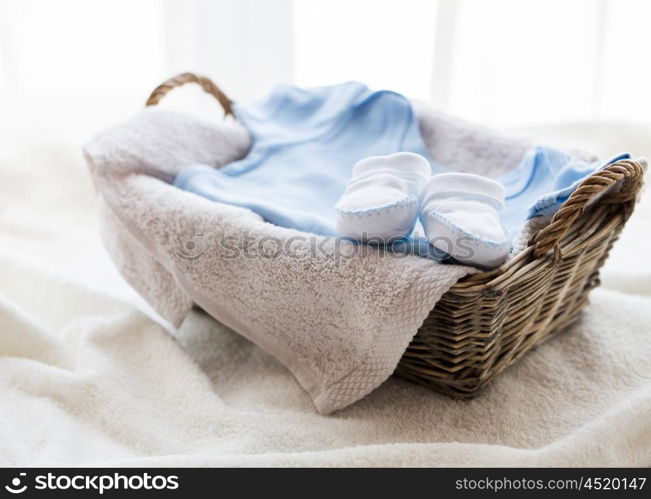 clothing, babyhood, motherhood and object concept - close up of white baby bootees with pile of clothes and towel for newborn boy in basket on table