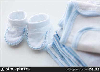 clothing, babyhood, motherhood and object concept - close up of white baby bootees with pile of clothes for newborn boy