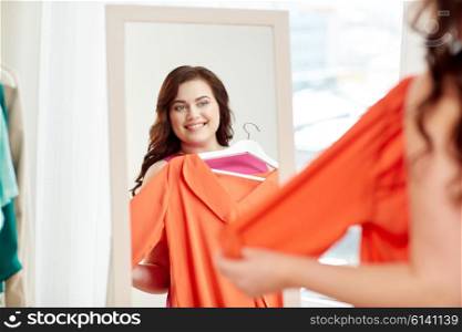 clothing, wardrobe, fashion, style and people concept - happy plus size woman with shirt on hanger at mirror at home