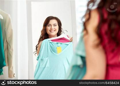 clothing, wardrobe, fashion, style and people concept - happy plus size woman with shirt on hanger at mirror at home. happy plus size woman with shirt at mirror