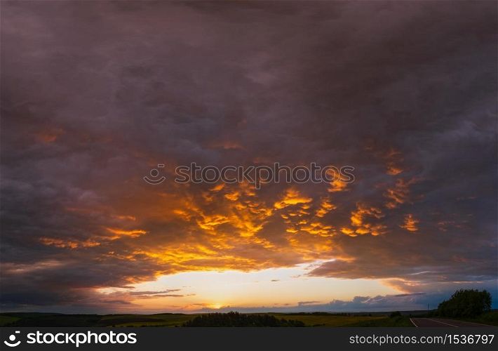 Cloudy sunset evening sky above spring rapeseed yellow fields, road and rural hills. Natural seasonal, weather, climate, countryside beauty concept and background scene.