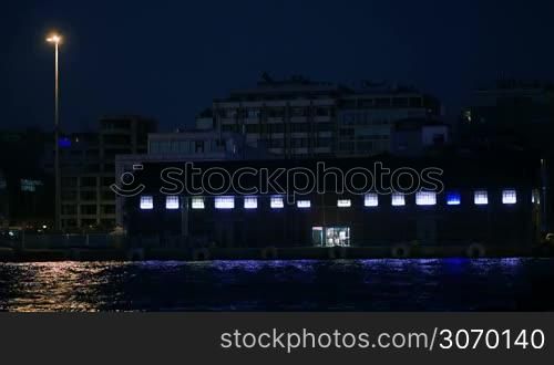 Coastal area on summer resort at night. Hotels in background and small illuminated building by the sea with blue light reflecting in water