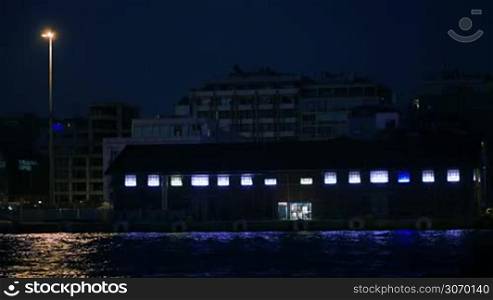 Coastal area on summer resort at night. Hotels in background and small illuminated building by the sea with blue light reflecting in water