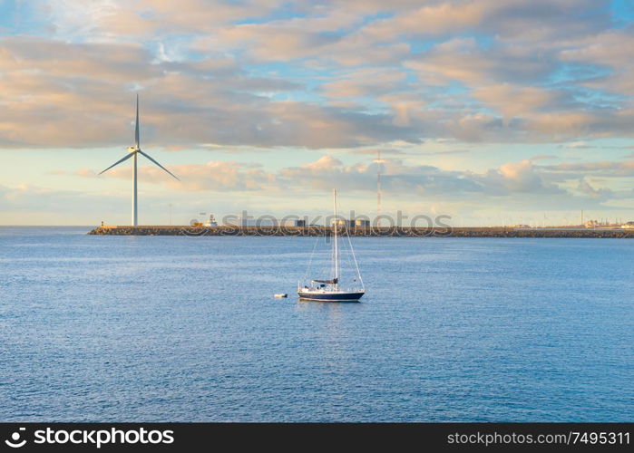 Coastal landscape with calm bay, breakwater, windmill and yacht at sunset