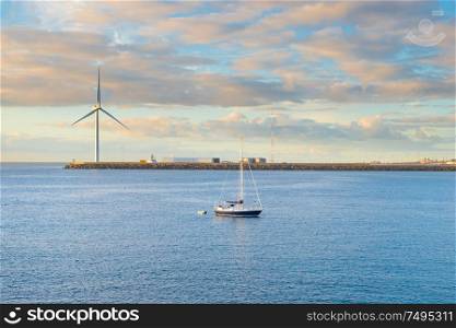 Coastal landscape with calm bay, breakwater, windmill and yacht at sunset