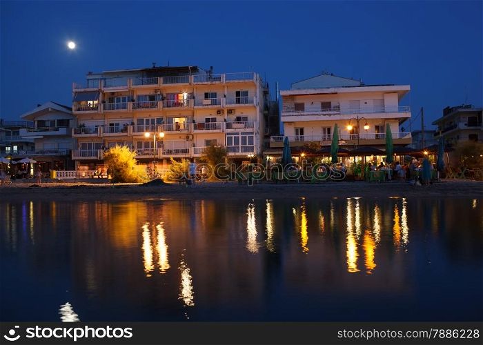 Coastal summer resort at night with outdoor cafe, hotel and people enjoying their vacation. Electric lights reflecting in water