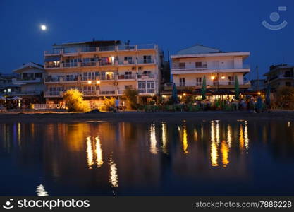 Coastal summer resort at night with outdoor cafe, hotel and people enjoying their vacation. Electric lights reflecting in water