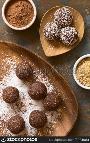 Coconut rum balls being covered with grated coconut on wooden plate, ingredients (cocoa powder, cookies) on the side, photographed overhead on slate with natural light