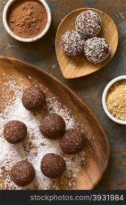 Coconut rum balls being covered with grated coconut on wooden plate, ingredients (cocoa powder, cookies) on the side, photographed overhead on slate with natural light