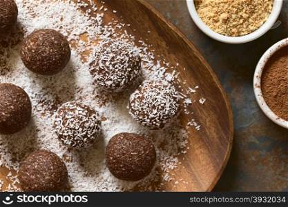 Coconut rum balls being covered with grated coconut on wooden plate, ingredients (cocoa powder, ground cookies) on the side, photographed overhead with natural light (Selective Focus, Focus on the top of the balls)