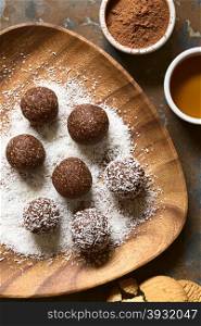 Coconut rum balls being covered with grated coconut on wooden plate, ingredients (cocoa powder, honey, cookies) on the side, photographed overhead on slate with natural light