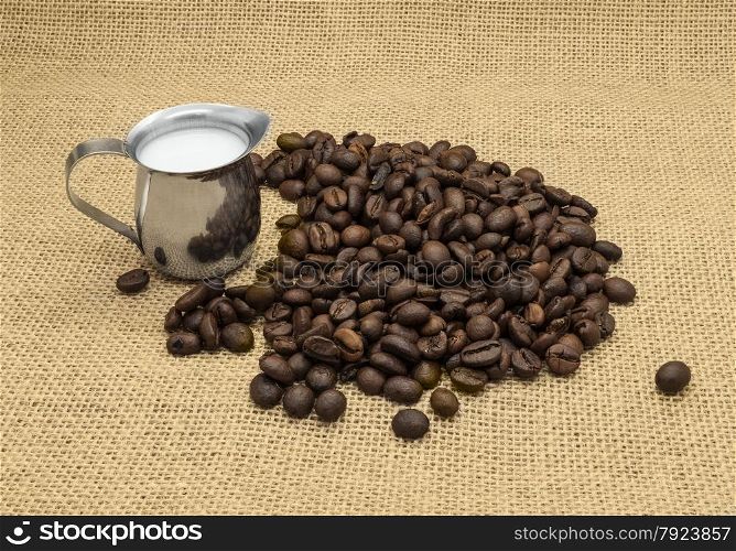 Coffee beans and milk jug on burlap background.