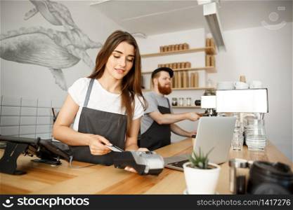 Coffee Business Concept - Beautiful female barista giving payment service for customer with credit card and smiling while working at the bar counter in modern coffee shop.