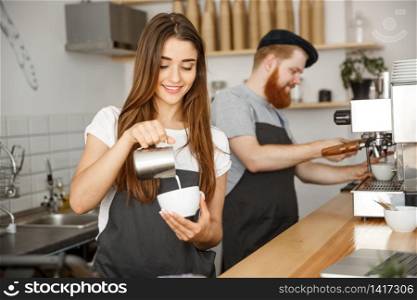 Coffee Business Concept - close-up lady barista in apron preparing and pouring milk into hot cup while standing at cafe.