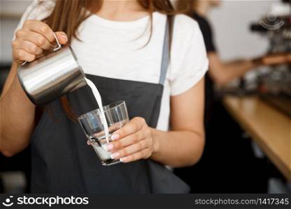 Coffee Business Concept - close-up lady barista in apron preparing and pouring milk in glass cup while standing at cafe.