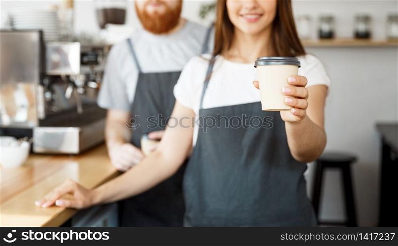 Coffee Business Concept - Positive young bearded man and beautiful attractive lady barista couple giving take away cup of coffee to custome at the modern coffee shop