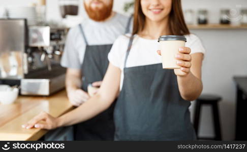 Coffee Business Concept - Positive young bearded man and beautiful attractive lady barista couple giving take away cup of coffee to custome at the modern coffee shop